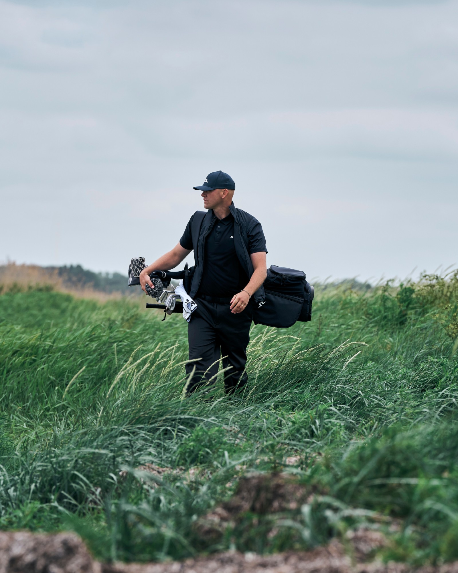 Golfer carrying a J.LINDEBERG bag through tall coastal grass