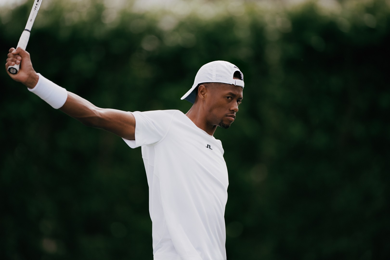 Tennis player in white stretching with a racket against a green hedge