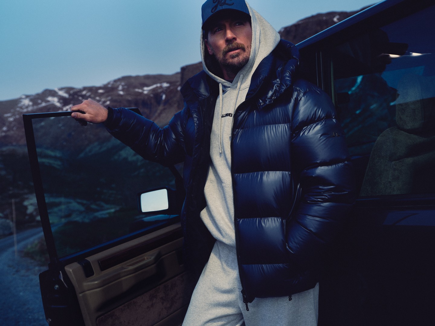 Close-up of a man in a navy puffer jacket holding an open car door at dusk