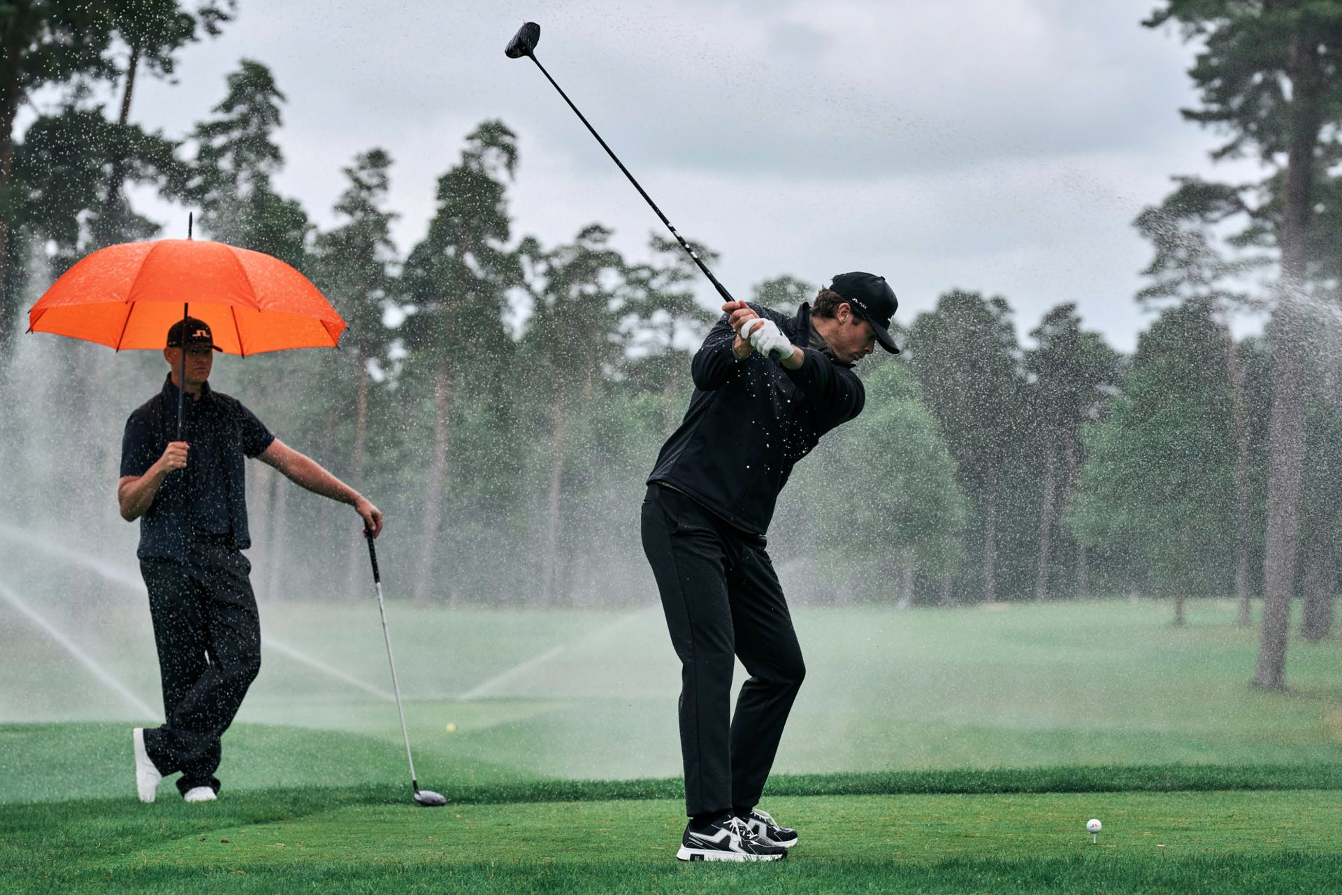 Golfer teeing off in the spray of course sprinklers while another waits under an orange umbrella