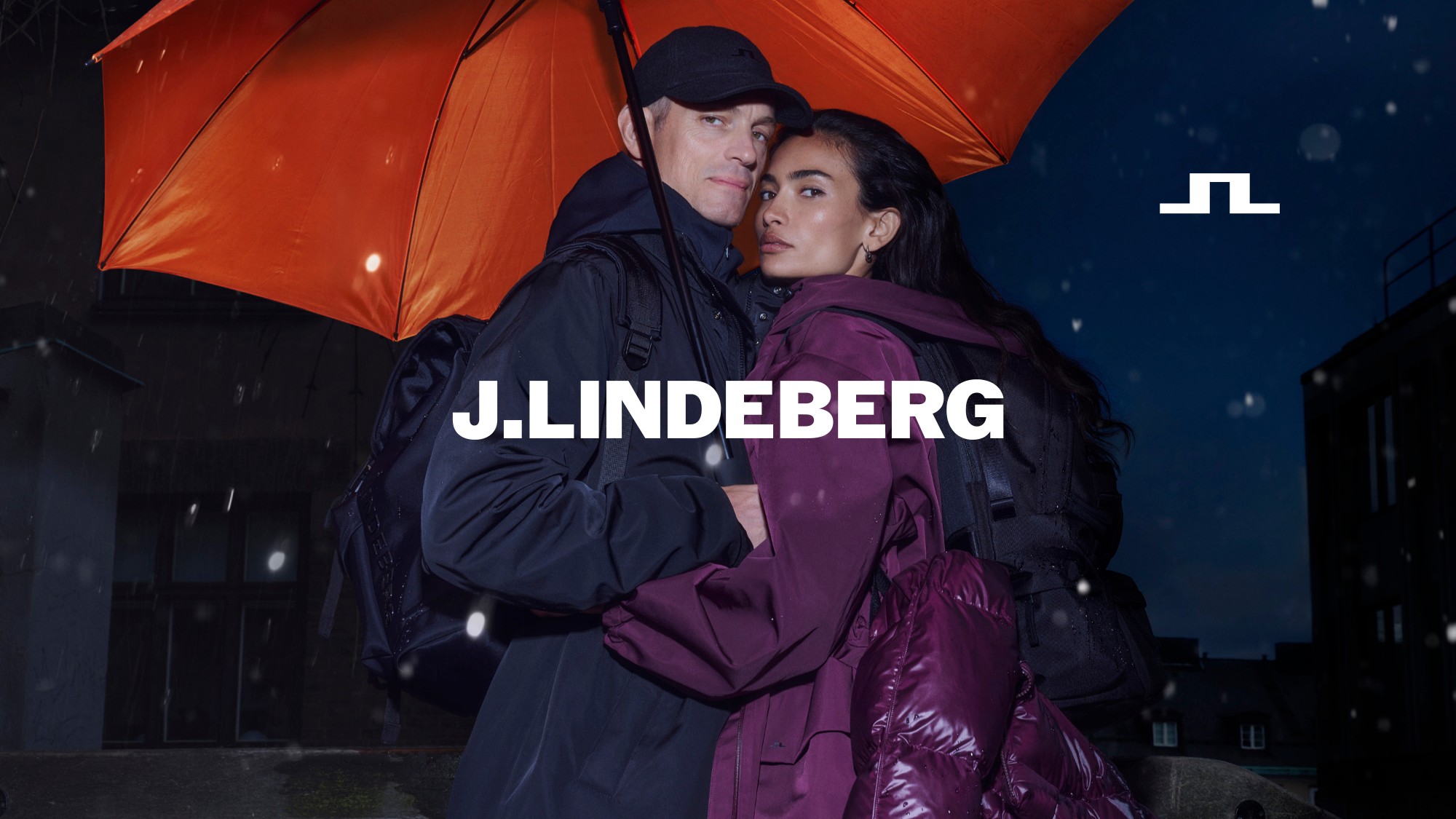 Joel Kinnaman and Kelly Gale under an orange umbrella at night, with the J.LINDEBERG logo