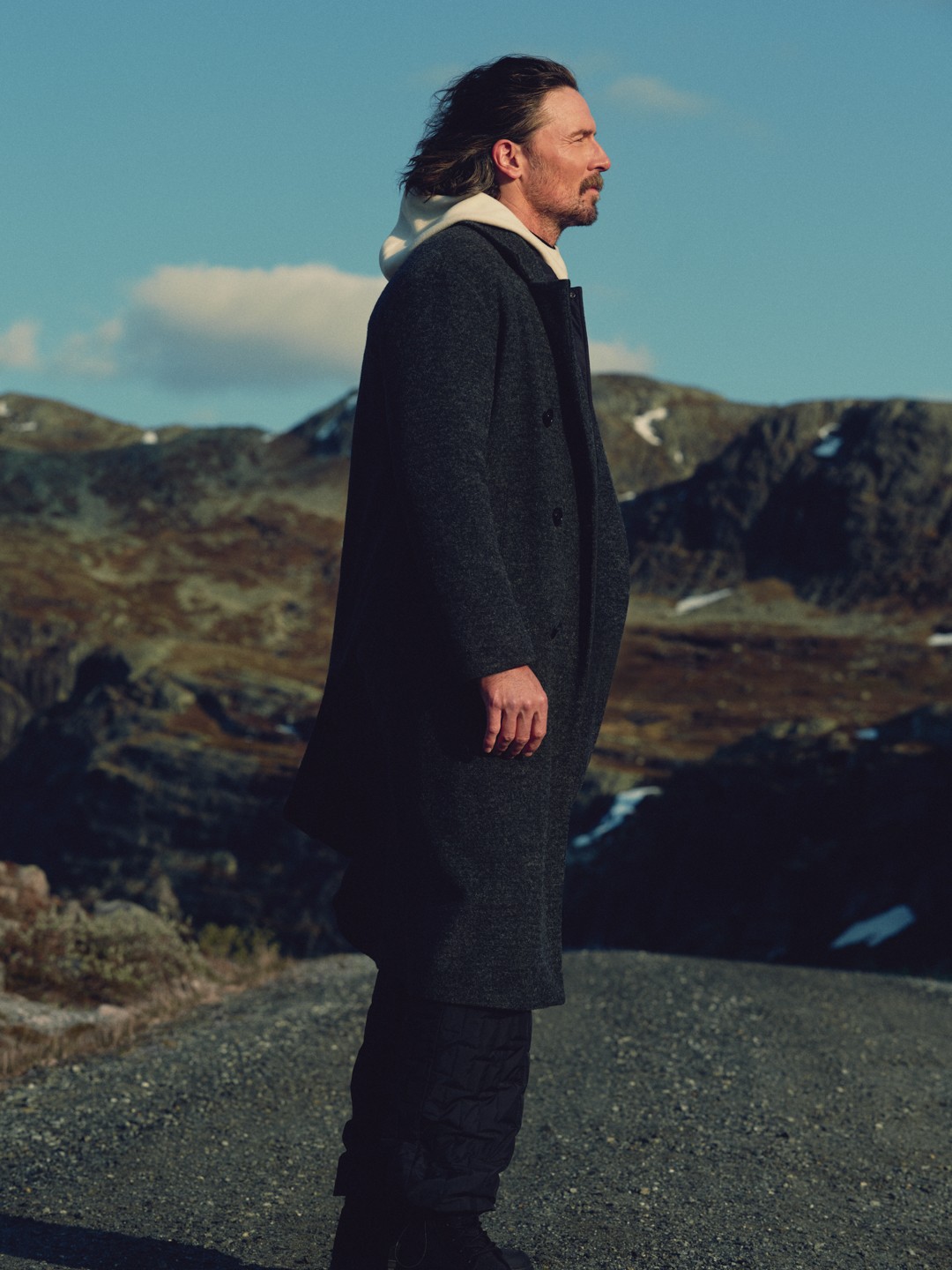 Man in a long charcoal coat standing on a mountain road in profile